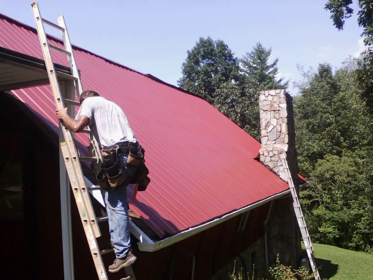Roofer on a ladder beside a red metal roof and stone chimney — Soffit & Fascia Repair by Roofing Brothers in Amity
